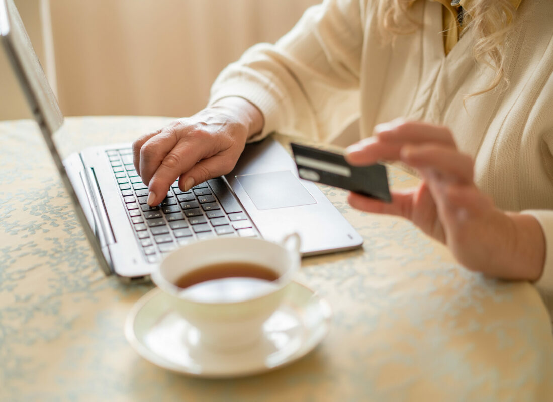 Selective Focus Of A Stylish Laptop On Which A Does Online Shopping. Blurred Cup Of Tea And Credit Card In Woman's Hand On The Foreground