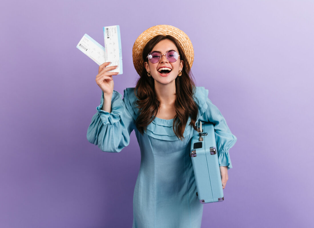 Dark-haired girl with glasses and straw hat holds tickets and blue suitcase. Portrait of traveler in cute retro dress on lilac background